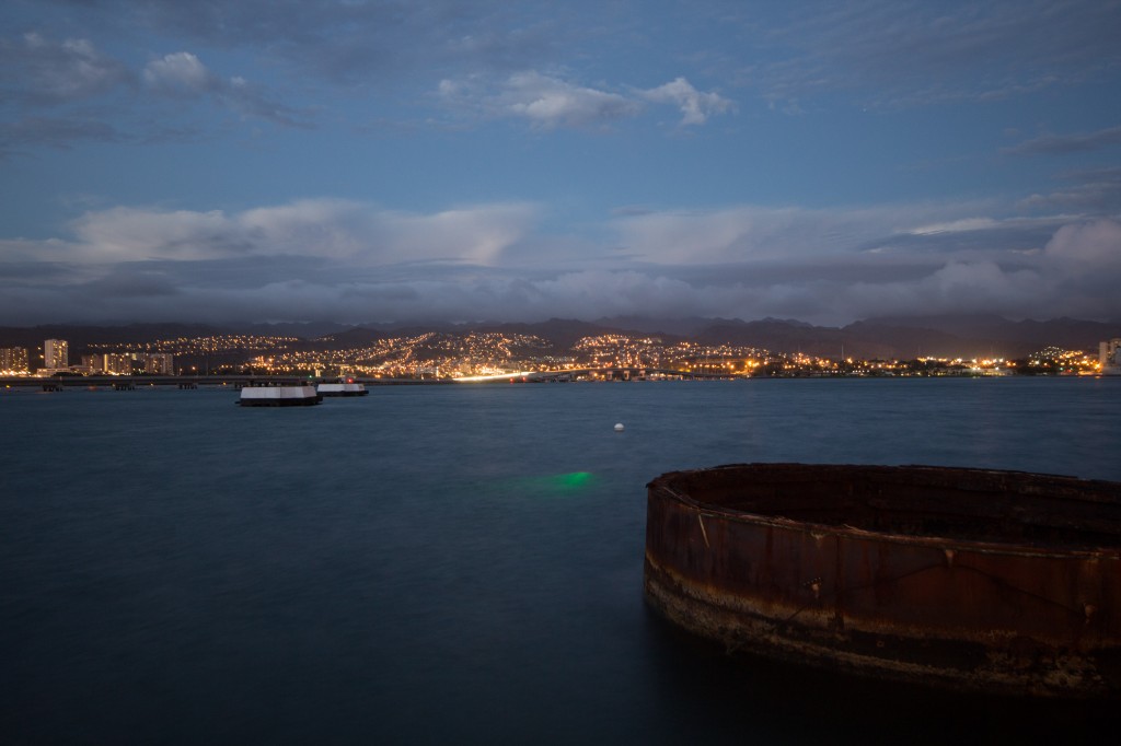 A view of Pearl Harbor, with the top of the USS Arizona in the foreground, and the green light of the underwater laser scanner glowing in the water.
